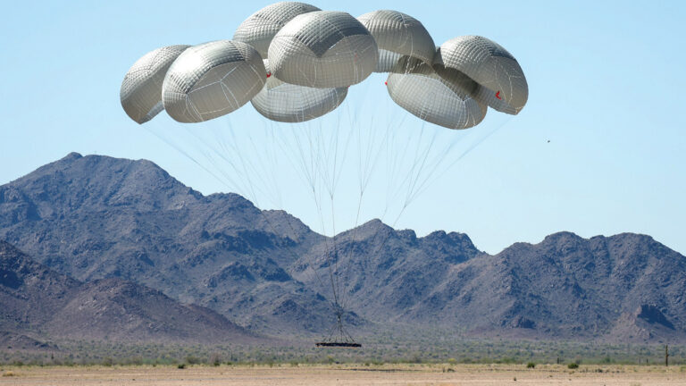 A group of six parachutes descends with a payload over a desert landscape, with rugged mountains in the background under a clear blue sky.