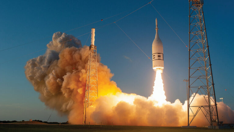 A rocket launches vertically amid a cloud of smoke and flames, flanked by two tall support towers against a clear blue sky.