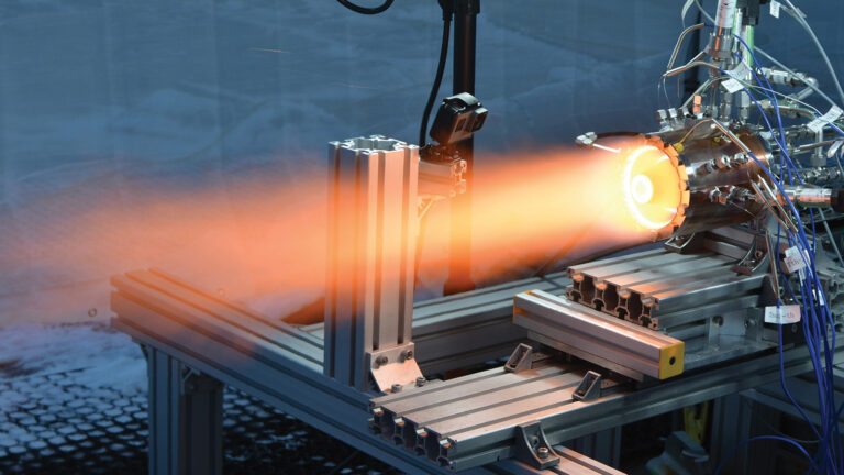 An industrial setup showcasing a scientific experiment with a device emitting a bright orange flame or light, surrounded by various cables and metal components.
