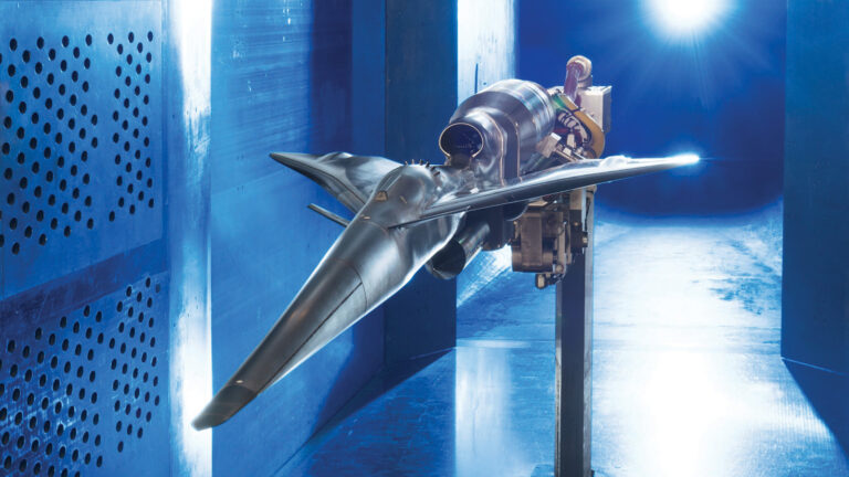 A futuristic aircraft model mounted on a testing rig is positioned inside a blue wind tunnel with light beams illuminating from the back.