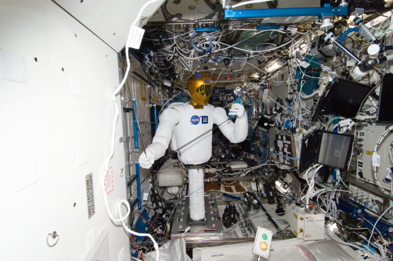 Robonaut 2, a humanoid robot with a gold visor, works inside the cluttered interior of the International Space Station, surrounded by various wires and equipment.