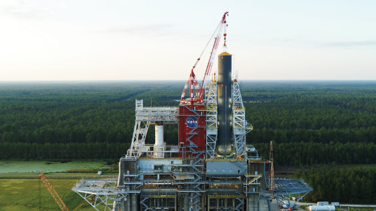 Aerial view of a NASA rocket on a launch pad surrounded by a forested area. Multiple cranes and scaffolding are seen around the structure under a clear sky.