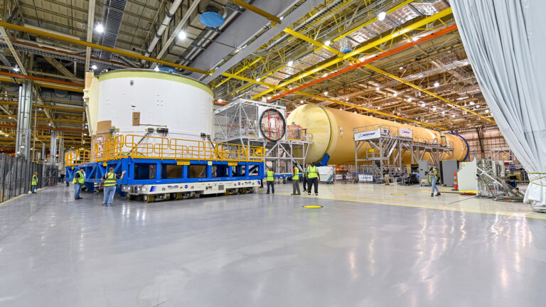 Space exploration focuses on asteroids Engineers and workers inside a large industrial facility assembling sections of a rocket or space vehicle with various equipment and machinery visible around.