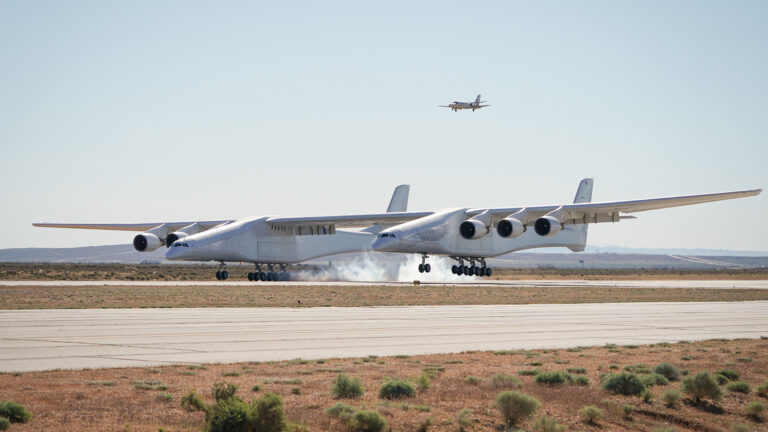 A large, six-engine aircraft touches down on a runway, with smoke visible from its landing gear. Another plane is seen flying in the background. The runway is surrounded by sparse vegetation.