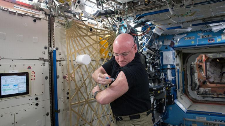 An astronaut administers a shot to himself while inside the International Space Station, surrounded by various equipment and panels.