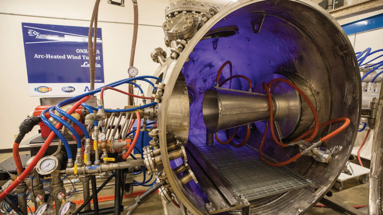 Hypersonics research accelerates across 3 continents Large industrial wind tunnel apparatus with various connected tubes, pipes, and gauges, displaying a conical structure inside. The equipment is metallic and partially illuminated with blue lighting.
