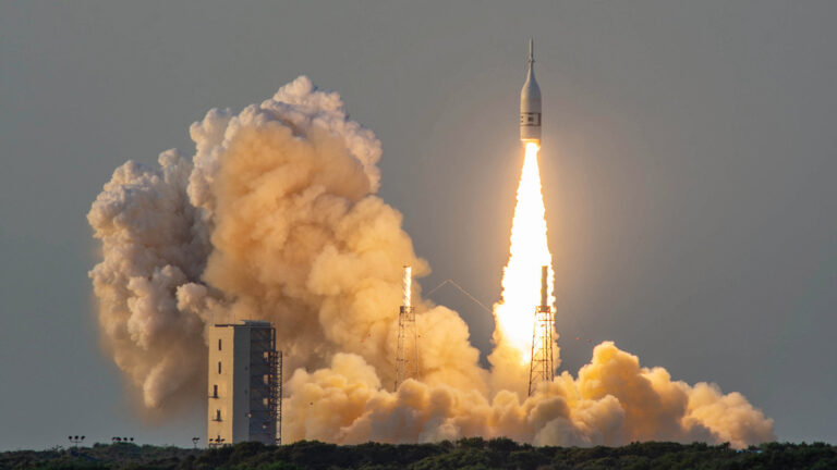 A rocket launches into the sky, leaving a trail of smoke and flames behind it as it ascends from the launch pad, with a large plume of smoke billowing at the base.