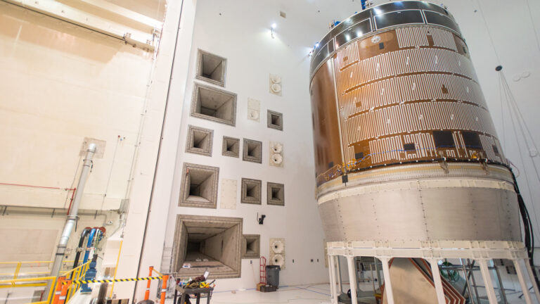 A large cylindrical spacecraft component is housed in a clean, white industrial facility with ventilation openings on the wall and various equipment and barriers surrounding it.