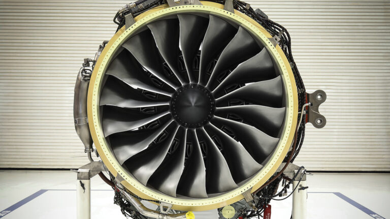 Close-up view of the front of an airplane jet engine showcasing multiple fan blades arranged in a circular pattern. The engine is mounted on a stand in a workshop environment.
