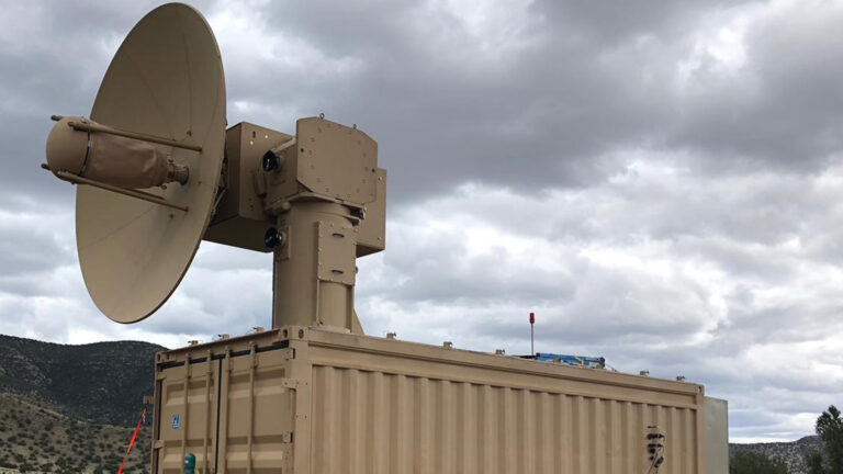 A large, tan-colored military communication satellite dish is mounted on a container in an outdoor setting with a cloudy sky.