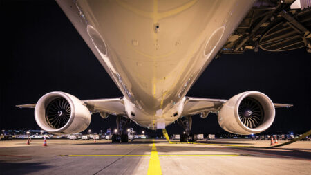 A close-up view of the underside of an airplane with its two engines visible, parked at an airport at night. The aircraft is connected to a jet bridge, and ground support equipment is in the background.
