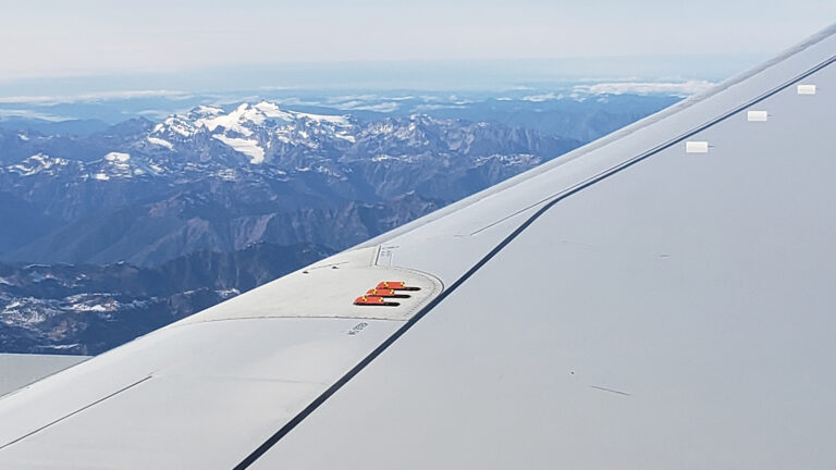 A view of snow-capped mountains from an airplane window, with the aircraft's wing prominently in the foreground under a clear sky.