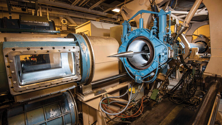 Image of an industrial machine with a large blue circular component and a metallic cone-shaped object inside, surrounded by various tubes, pipes, and wiring.