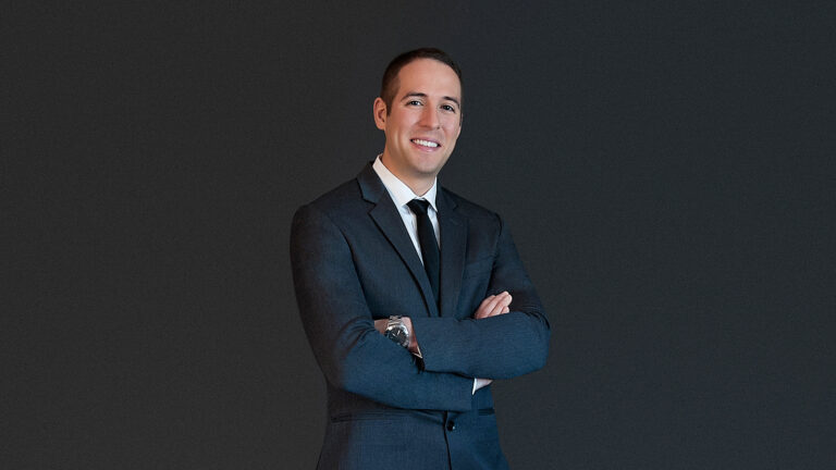 A man in a suit with a tie stands against a grey background, smiling with his arms crossed.
