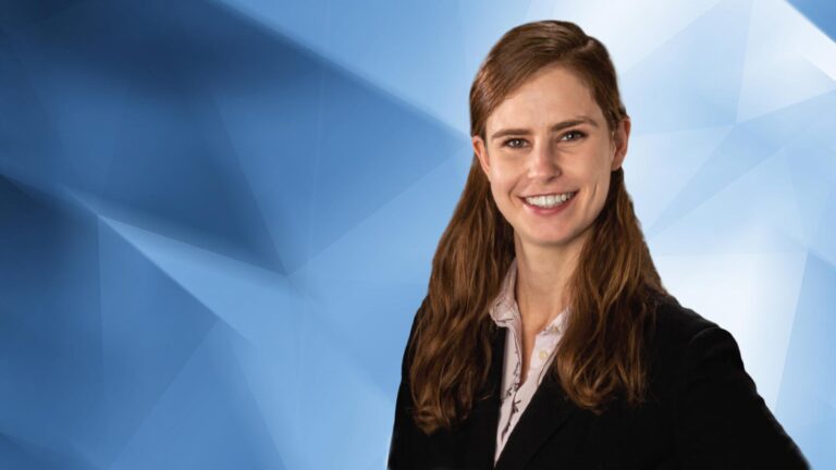 A woman with long brown hair, smiling and wearing a black blazer over a light-colored blouse, stands in front of a blue geometric background.