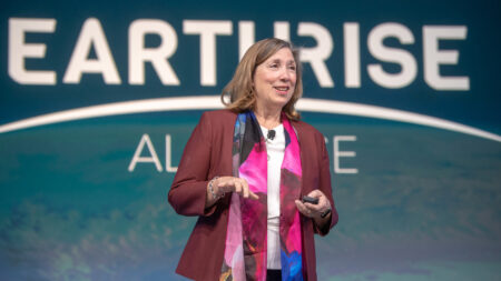 A woman in a maroon blazer and colorful scarf speaks on stage in front of a backdrop that says "EARTHRISE.