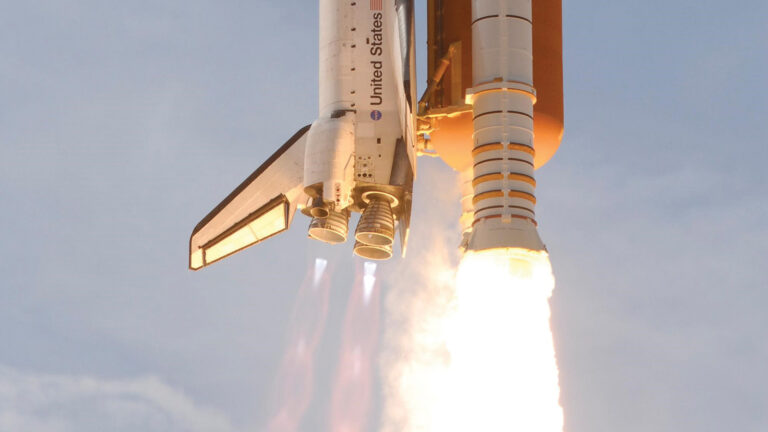 A space shuttle is launching with engines firing, emitting bright flames and smoke against a clear sky.