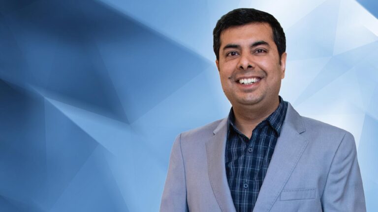 A man in a grey blazer and checkered shirt smiles, standing in front of a blue geometric background.