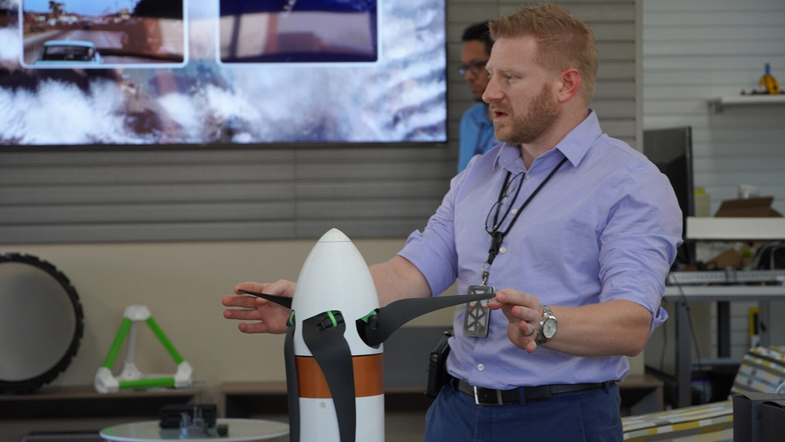 A man in a light blue shirt and black lanyard is gesturing towards a model rocket while presenting in an indoor setting with a TV screen in the background.