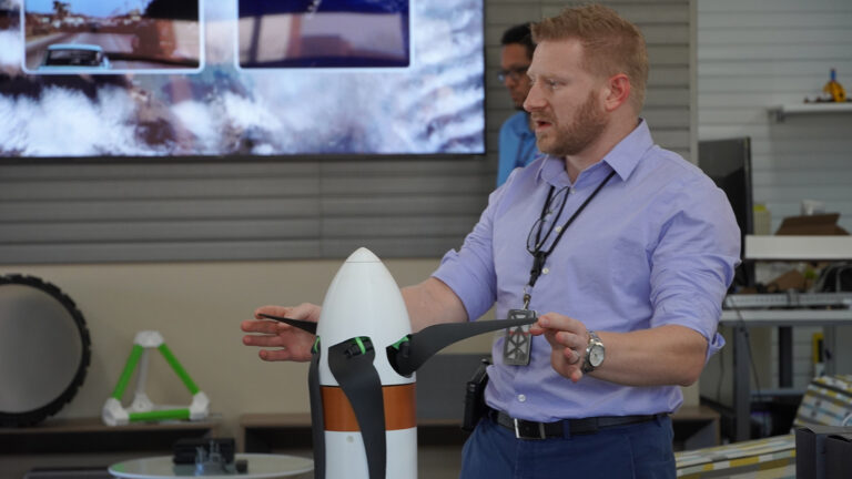A man in a light blue shirt and black lanyard is gesturing towards a model rocket while presenting in an indoor setting with a TV screen in the background.