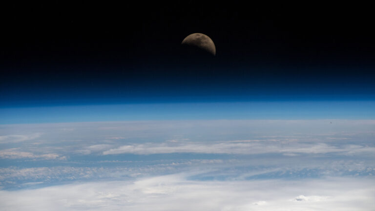 A view from space of Earth's horizon with a partially shadowed moon in the background against a dark sky.