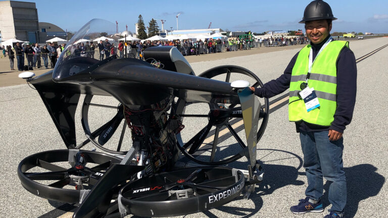 A person wearing a safety vest and helmet stands next to an experimental VTOL aircraft on a runway, with a crowd and tents visible in the background.