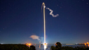 A rocket launches into the night sky, leaving a bright curved trail of light, with stars visible in the background and reflections on a water body below.