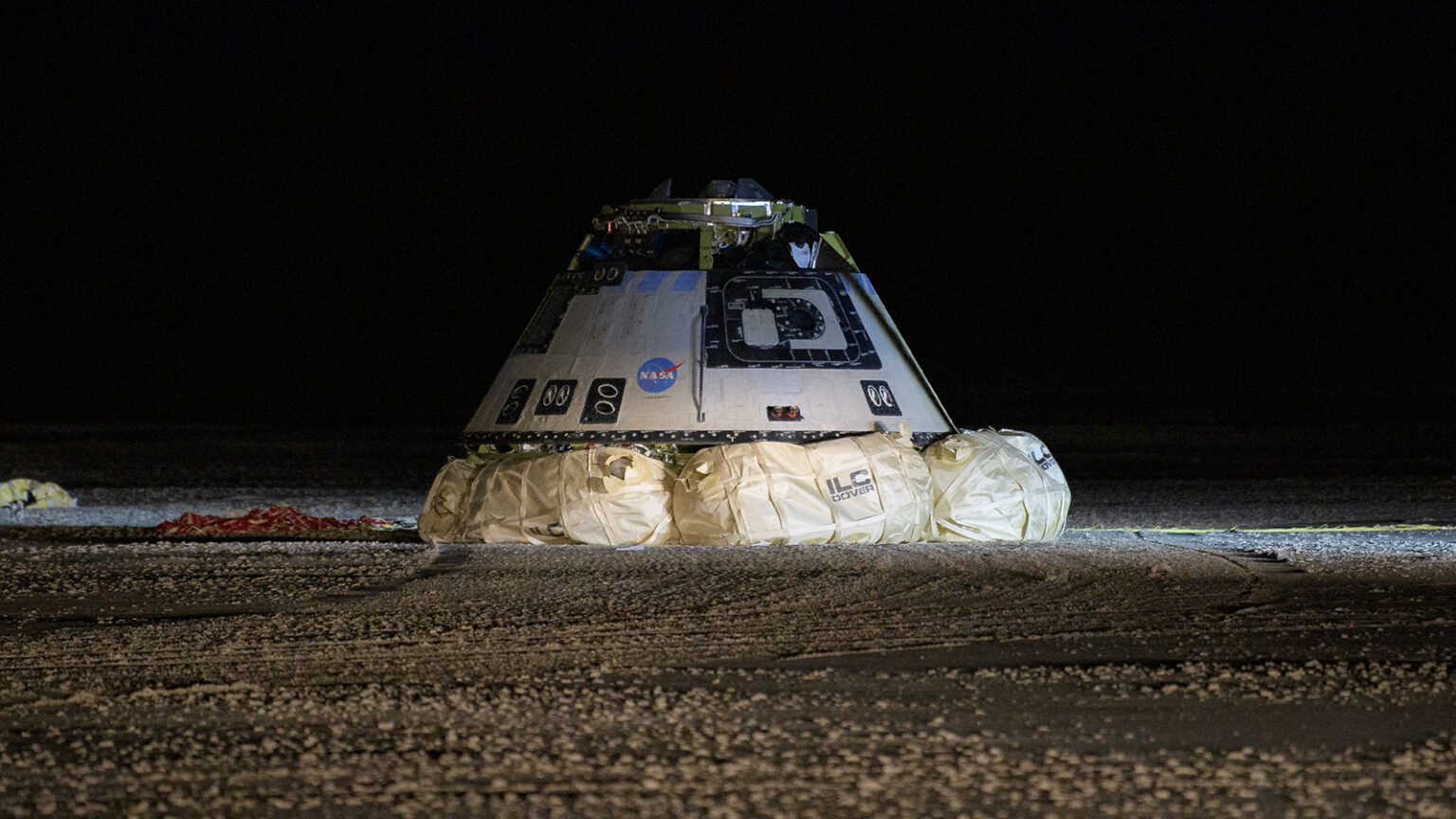A space capsule sits on the ground at night, surrounded by inflated airbags used for landing. Numbers and letters adorn the capsule, with visible branding from NASA and other organizations.