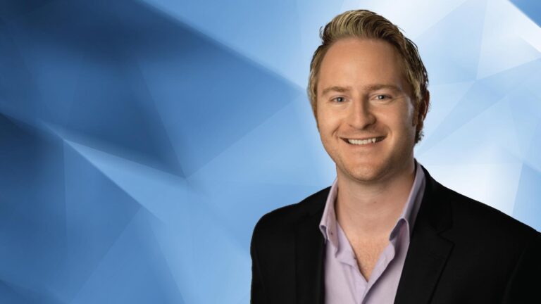 A smiling person with short blonde hair wearing a black blazer over a light purple shirt, standing against a blue geometric background.
