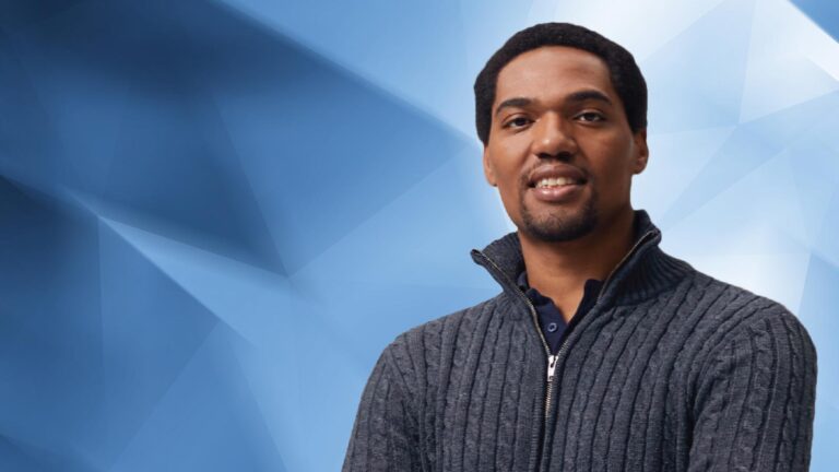 A man wearing a dark zip-up sweater stands against a blue geometric background, smiling slightly at the camera.