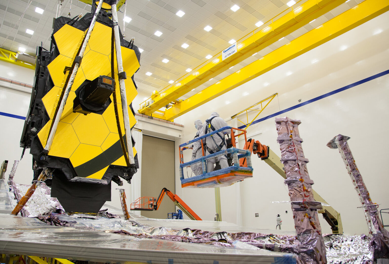 Engineers work on a large, segmented golden mirror of a space telescope in a clean room, using a cherry picker and surrounded by reflective materials.