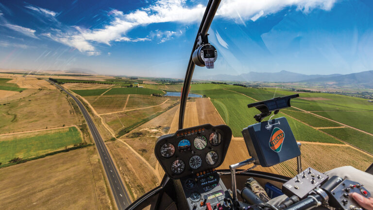 View from inside a helicopter cockpit flying over a landscape with fields, roads, and distant mountains under a partly cloudy sky.