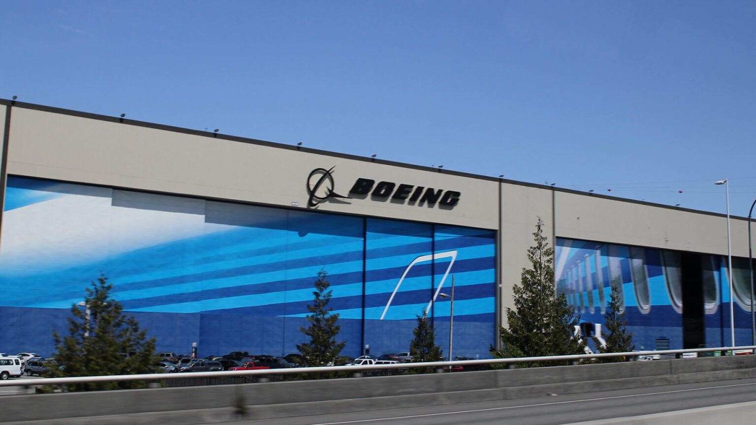 A large industrial building with the "Boeing" logo on it, featuring blue themed murals on its exterior, with parked cars in front and trees along the road leading up to it.