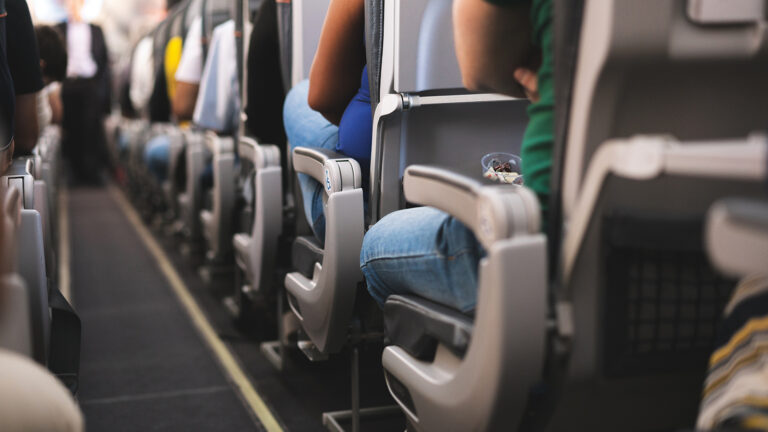 Passengers seated in rows inside an airplane; focus is on the middle section of the cabin, showing the back of seats and armrests.