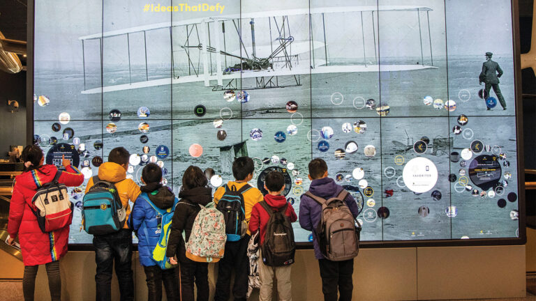 Children with backpacks stand in front of a large interactive display featuring an image of a historic airplane, engaging with the screen and its various digital icons.