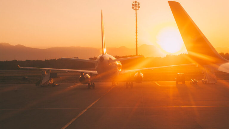 An airplane parked on an airport tarmac during sunset with mountains in the background and a distant light tower.