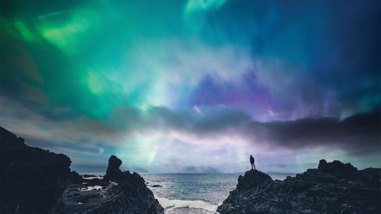 Person standing on a rocky shore, gazing at the Northern Lights illuminating the sky with vibrant green and purple hues above a calm sea.