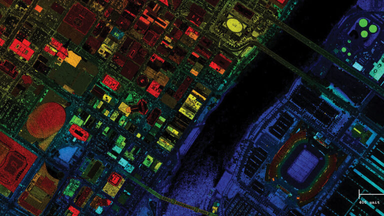 A colorful satellite image showing a city layout with various buildings, roads, and a large stadium. A river or water body divides the densely built area from the section with the stadium.