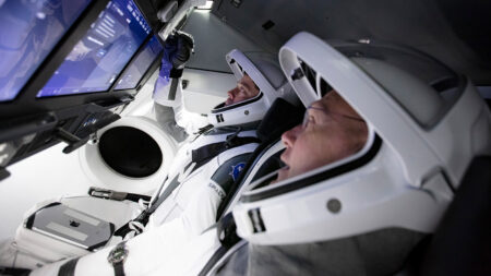 Two astronauts sit inside a spacecraft cockpit, using touchscreens on a control panel. Both are wearing white space suits and helmets.