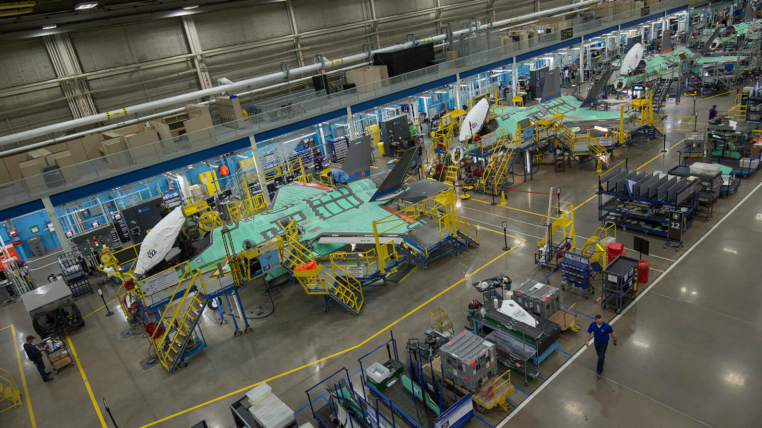 An aircraft production line in a factory with several partially assembled planes, various workstations, and workers engaged in different tasks.