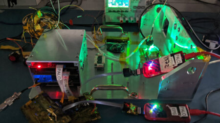 A cluttered electronics workstation with various components, wiring, and circuit boards illuminated by green and red lights. A metal case and labeled connectors are among the visible items.