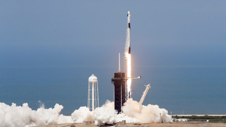A rocket launches into the sky from a seaside launch pad, leaving a trail of exhaust. A water tower is visible nearby.