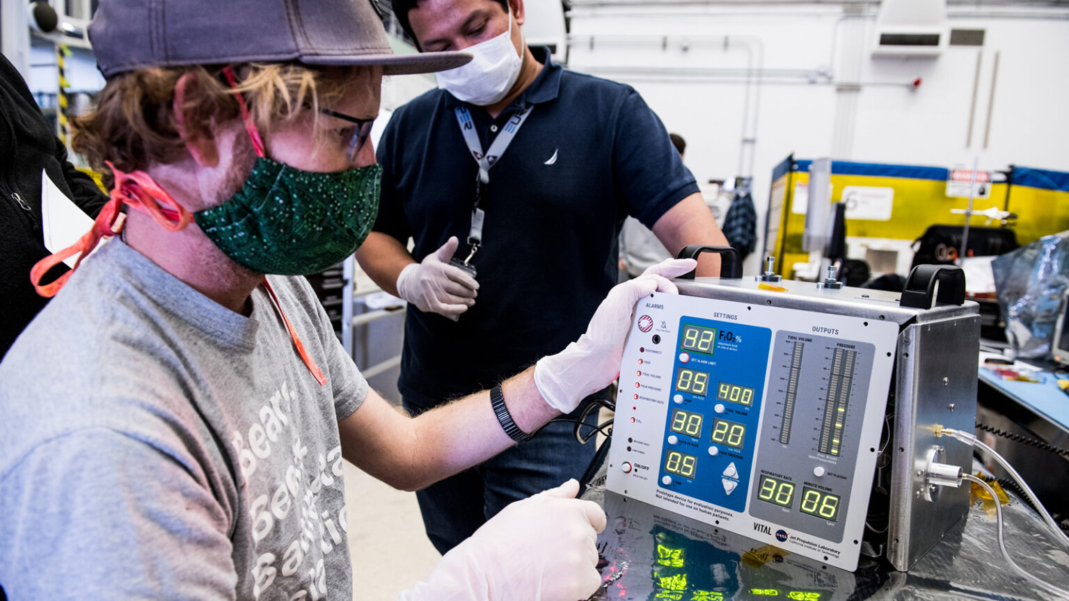 Two technicians in a lab work with a medical device. Both are wearing gloves, one is wearing a mask, and the other has a mask around his neck. The devices show measurement readings on a digital display.