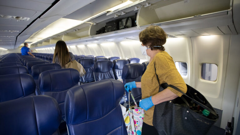 A woman wearing gloves places a bag in the overhead compartment of an airplane, while another passenger walks down the aisle.