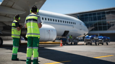 Two ground crew members in reflective uniforms and earmuffs stand near an airplane on the tarmac, with the terminal building in the background.
