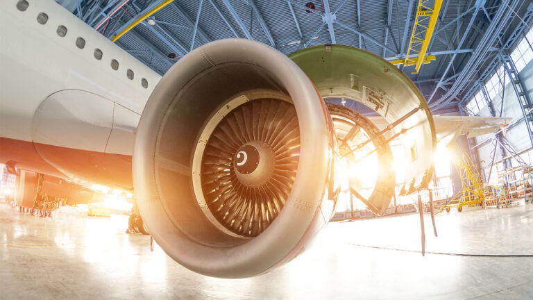 Close-up of an airplane engine in a hangar, with opened panels revealing internal components. The background shows structural elements and sunlight streaming through windows.