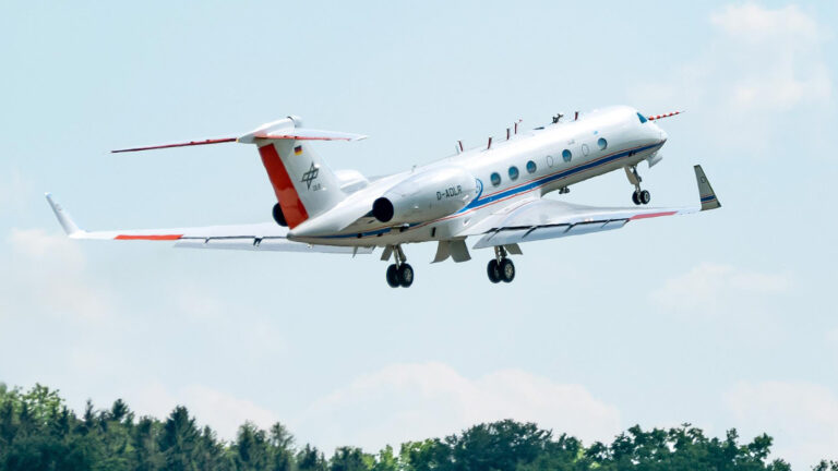 A white private jet with red and blue accents takes off from a runway, surrounded by a backdrop of trees and a clear sky.