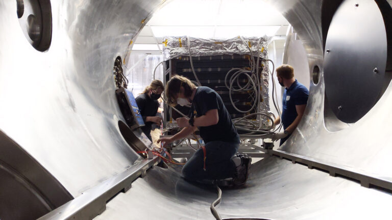 Three individuals work inside a large cylindrical metal structure, handling cables and tools. One person kneels in the center, while the others assist. The background reveals additional equipment.