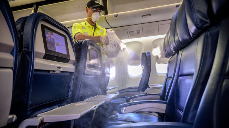 A person in protective gear disinfects the interior of an airplane, spraying cleaning solution on the seats and tray tables.