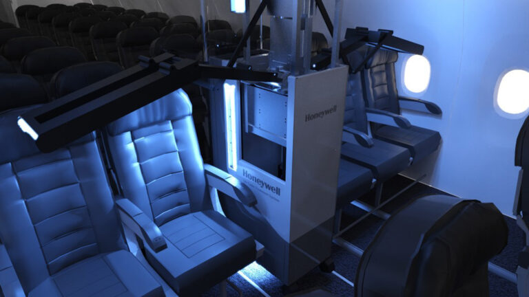 Image shows an airplane cabin undergoing disinfection by a Honeywell robotic system, with ultraviolet lights extended over rows of seats. The cabin is empty and illuminated by blue UV light.